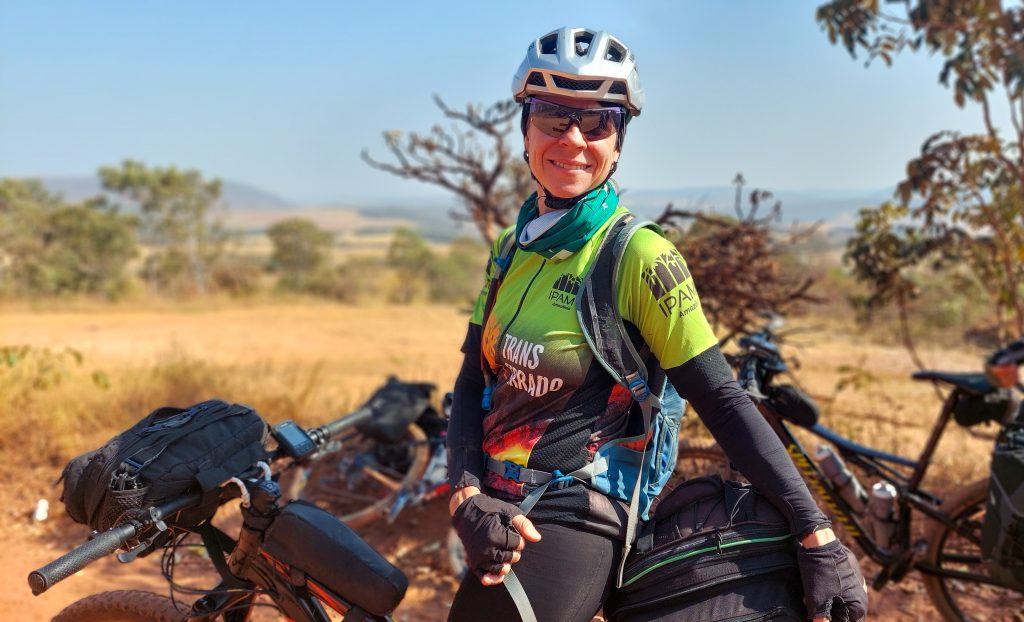 A foto mostra mulher sentada numa biciclicleta em meio a vegetação cerratense. Ela é a primeira mulhere no TransCerrado.
