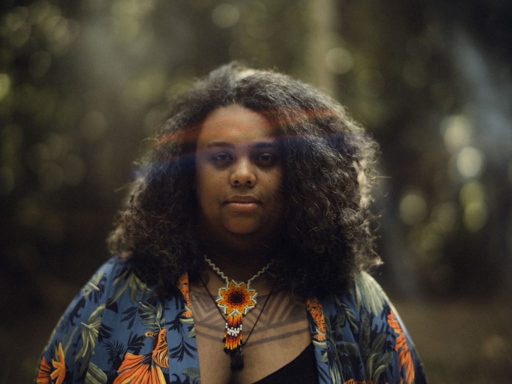 Retrato de Kamila Camilo. Uma mulher preta trajando uma camisa azul com detalhes de flores com pétalas laranjas. Ela usa um colar indígena no pescoço e um raio do sol repousa em sua cabeça. 