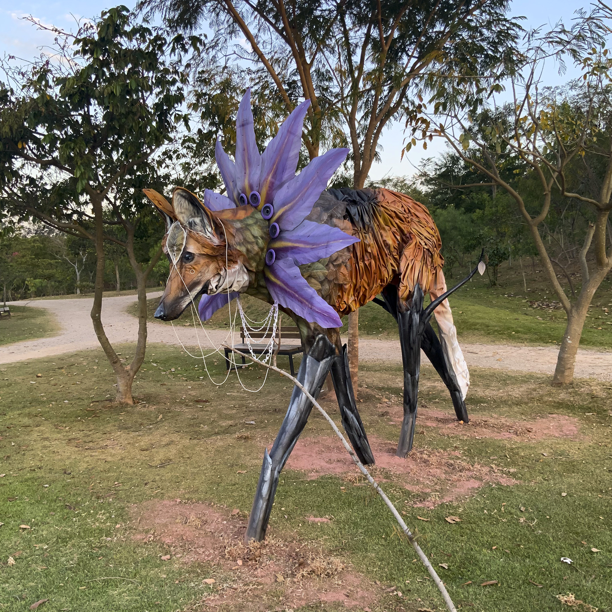 Escultura de lobo-guará feita com materiais descartados que representam ameaças ao animal, como lona de caminhão (colisão/atropelamento veicular) e barril de plástico (agrotóxicos ilegais). As patas representam arvores queimadas, como uma extensão da flora. A espécie percorre cerca de vinte quilômetros diários, espalhando sementes de frutas do qual se alimenta, como a fruta-do-lobo. A flor do fruto serve como coroa na obra.